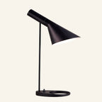 Matte black table lamp with tilting asymmetric cone shade, slim stem and oval ring base, Bauhaus-style metal task light on cream backdrop