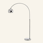 Polished chrome arc floor lamp with slender curving stem, hemispherical dome shade, and round mirrored base on cream studio backdrop