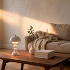 Lit white mushroom lamp on a walnut coffee table beside an olive sprig vase and stacked books, with linen sofa in warm evening light