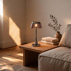 Cordless chrome bedside lamp glowing on an oak coffee table beside stacked books and an olive sprig, sunlit minimalist living room