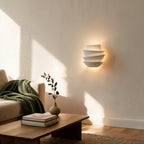 Petal wall sconce glows warmly in a sunlit living room above a wooden coffee table with olive sprig, books, and green throw
