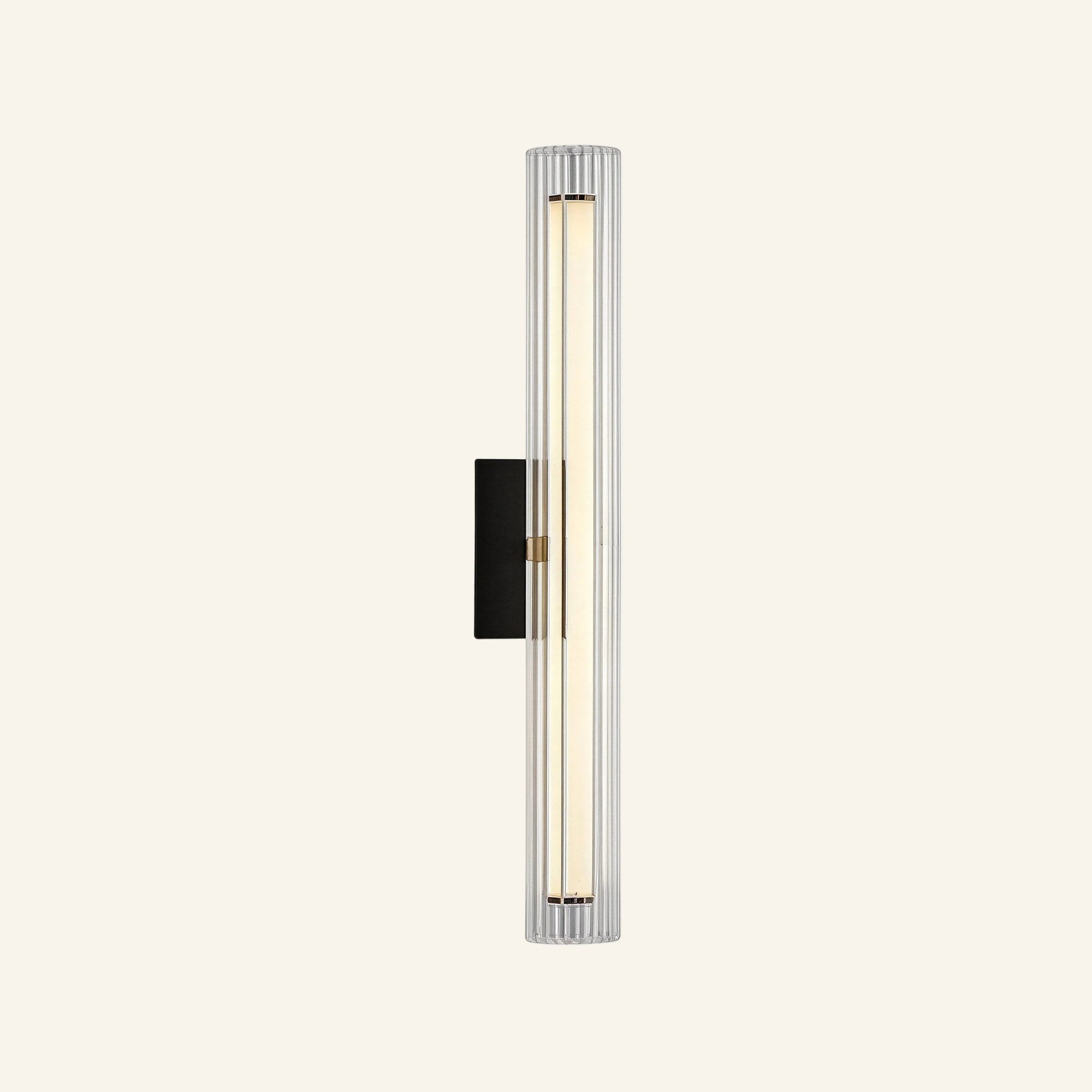 Slim vertical wall sconce with fluted clear glass column, matte black backplate, and brass center band housing a warm LED rod