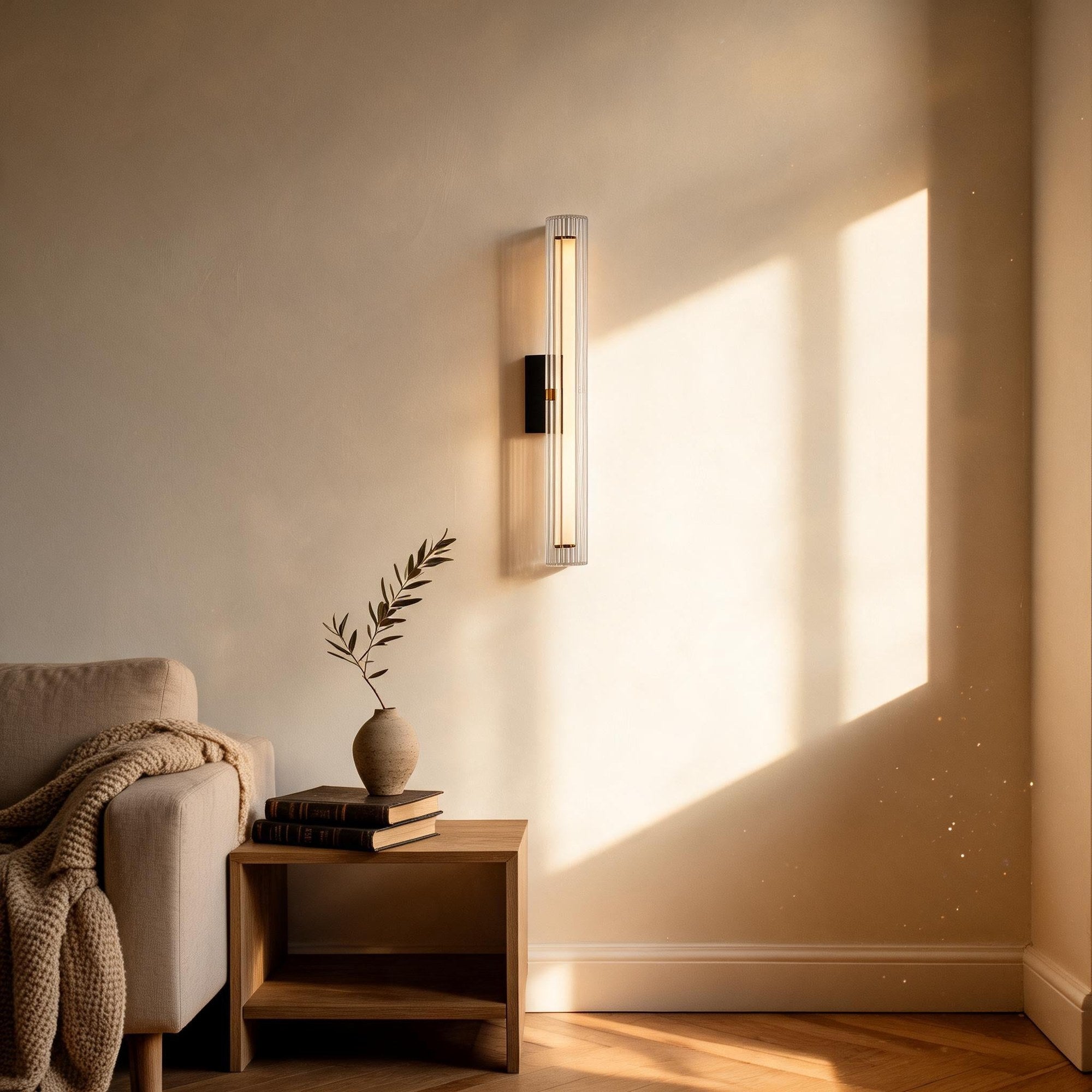 Fluted glass sconce mounted on a plaster wall above a wooden side table with books and an olive sprig in a stone vase, sunlight streaming across a reading nook