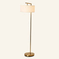 Slim brass floor lamp with cantilevered arm holding a white linen drum shade above a round weighted base on cream backdrop.