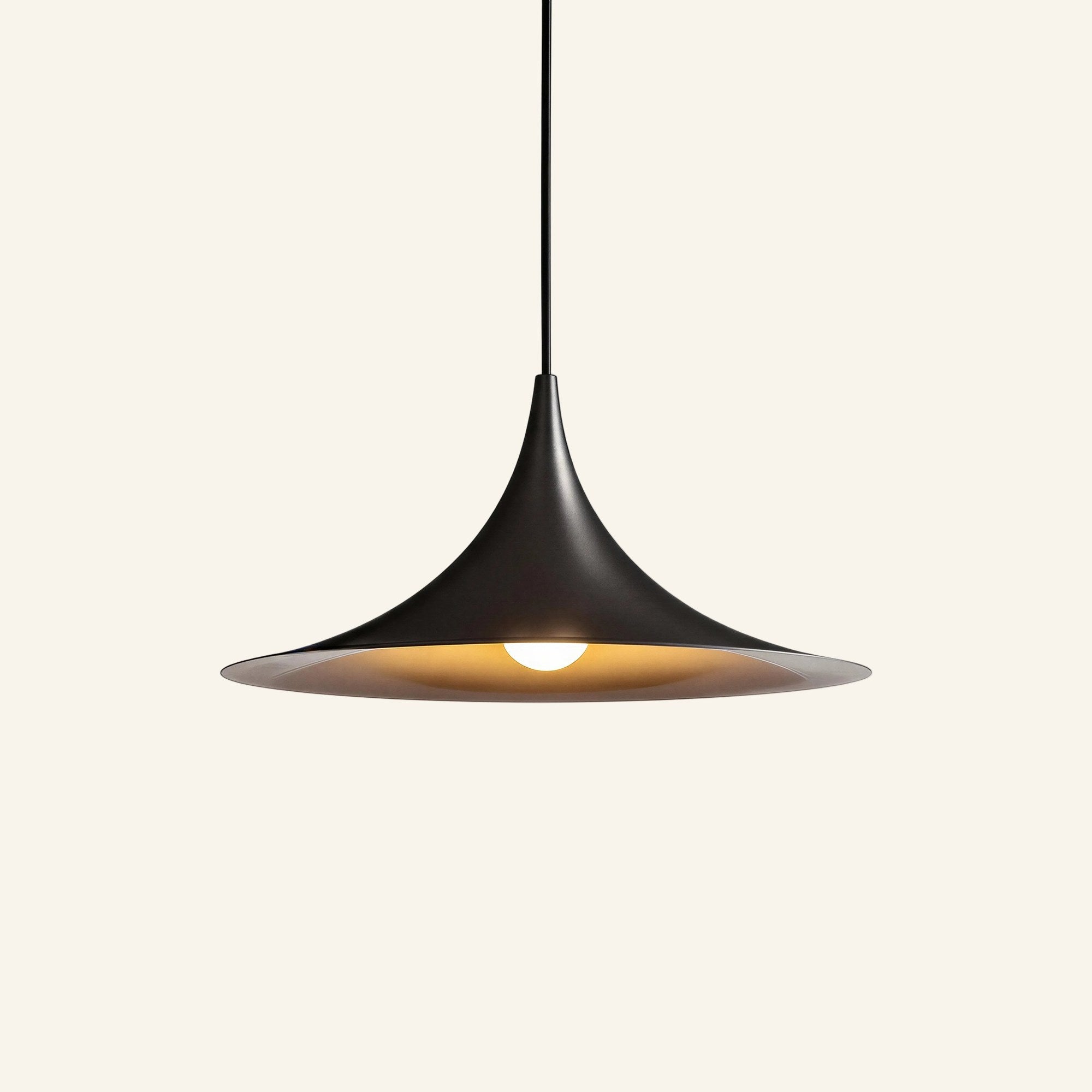 Matte black trumpet-shaped pendant lamp with wide flat brim and warm bulb glow, suspended from a black cord on cream studio backdrop.