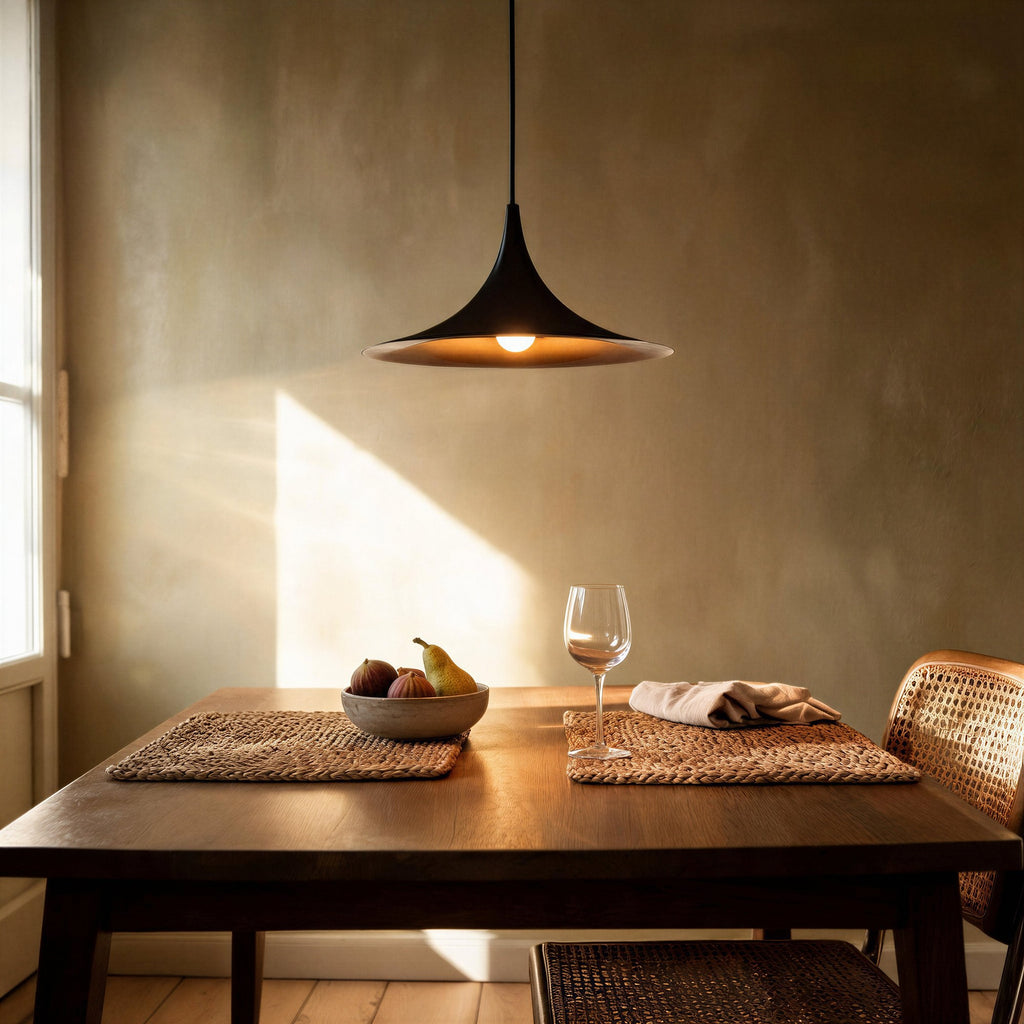 Black pendant lamp hanging above a wooden dining table set with figs, wine glass and rattan chair in a sunlit room.