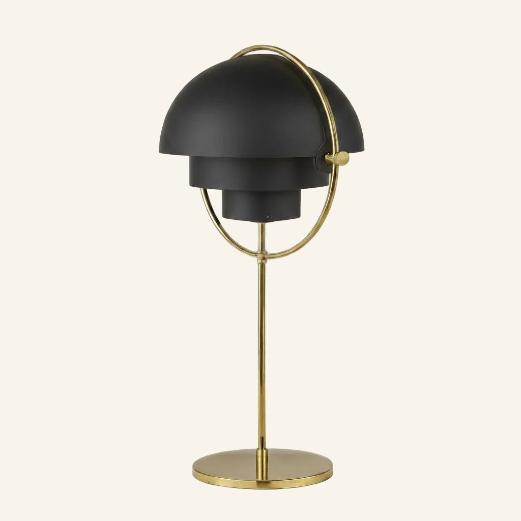 Matte black twin-dome shades rotate within a brass ring on a slim brass stalk and circular base, sculptural table lamp on neutral background.