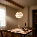 Petal pendant lamp glowing warmly above an oak dining table set with wine, figs and a rattan chair beneath a beamed ceiling