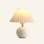 Illuminated table lamp with white pleated fabric shade, polished brass neck, and glossy spherical ceramic base on cream backdrop