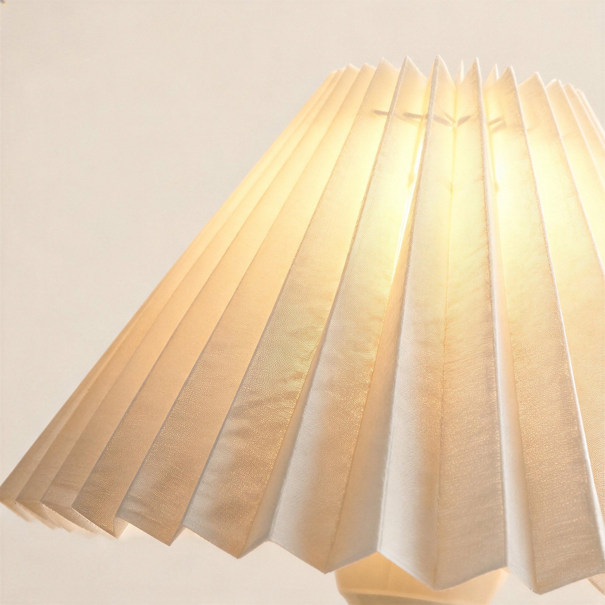 Close-up of the crisp accordion pleats on the ivory fabric shade, glowing warmly to reveal woven texture and brass-trimmed neck