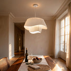 Pleated silk pendant lamp glowing above a wooden dining table with wine, fruit bowl and rattan chair in a sunlit room