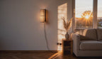 Cinematic video of a cylindrical linen wall sconce glowing warmly beside a sofa as sunset light streams through the window.