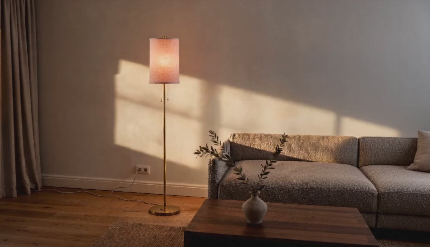 Cinematic video of a slim cream linen drum floor lamp glowing beside a textured sofa in a warm living room at dusk.