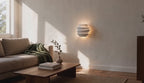 Cinematic video of a tiered matt white petal wall sconce glowing warmly above a sofa in a sunlit living room.
