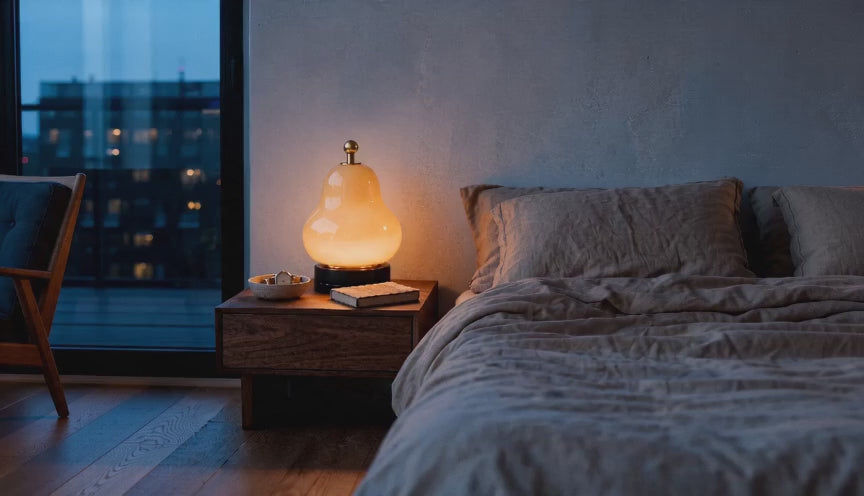 Cinematic bedroom video of a pear-shaped glass table lamp glowing warmly on a wooden nightstand beside linen bedding at dusk.