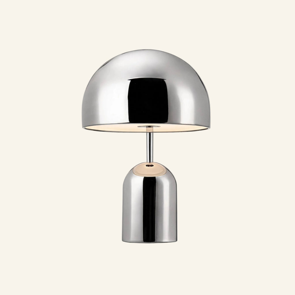 Polished chrome mushroom-shaped table lamp with domed shade and cylindrical base, glowing warmly on a cream studio backdrop