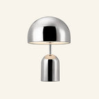 Polished chrome mushroom-shaped table lamp with domed shade and cylindrical base, glowing warmly on a cream studio backdrop