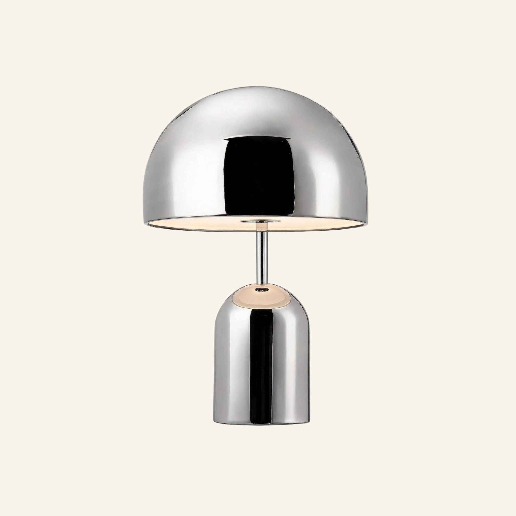 Polished chrome mushroom-shaped table lamp with domed shade and cylindrical base, glowing warmly on a cream studio backdrop