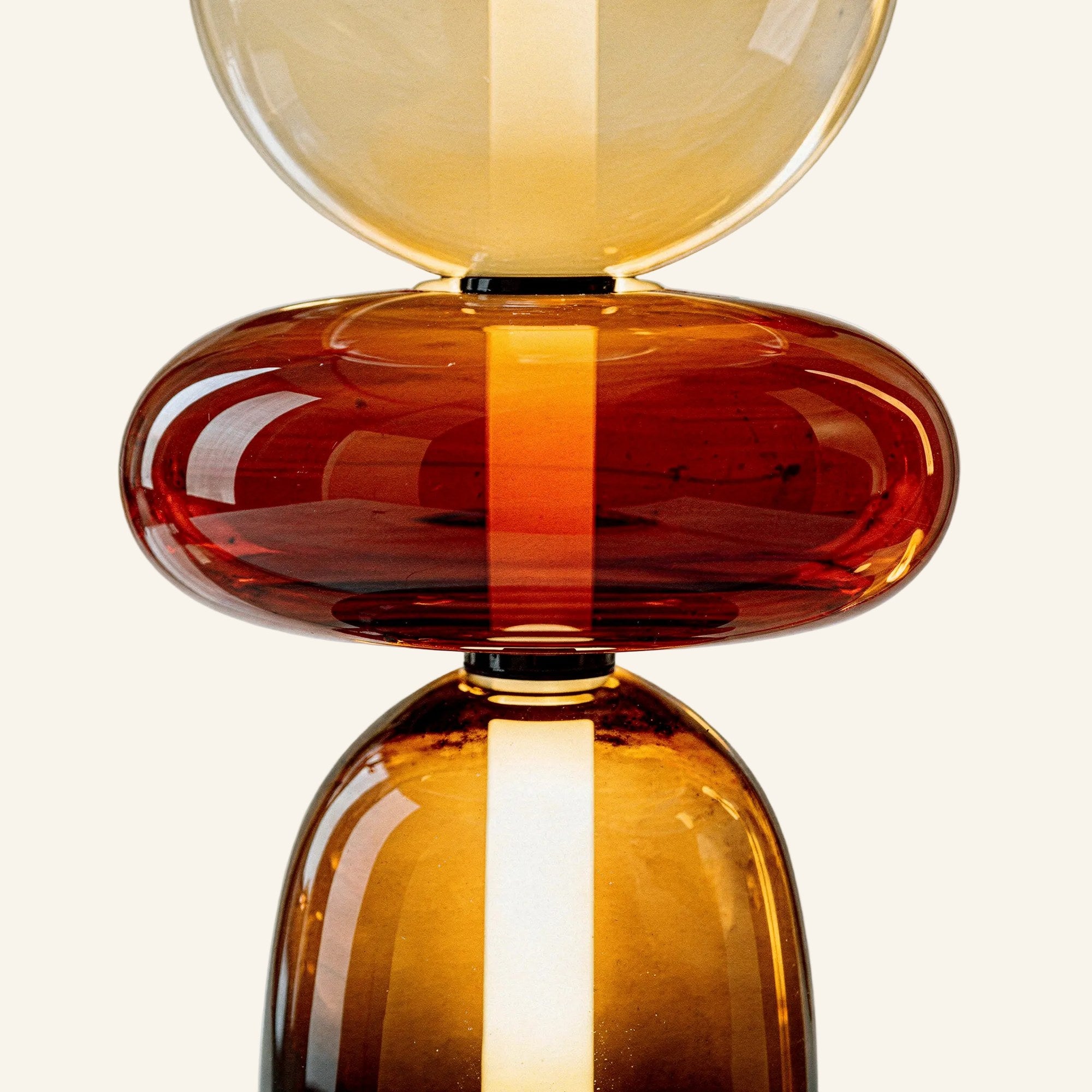 Close-up of hand-blown borosilicate glass pebbles in amber and cherry tones, internal light column glowing through the translucent column