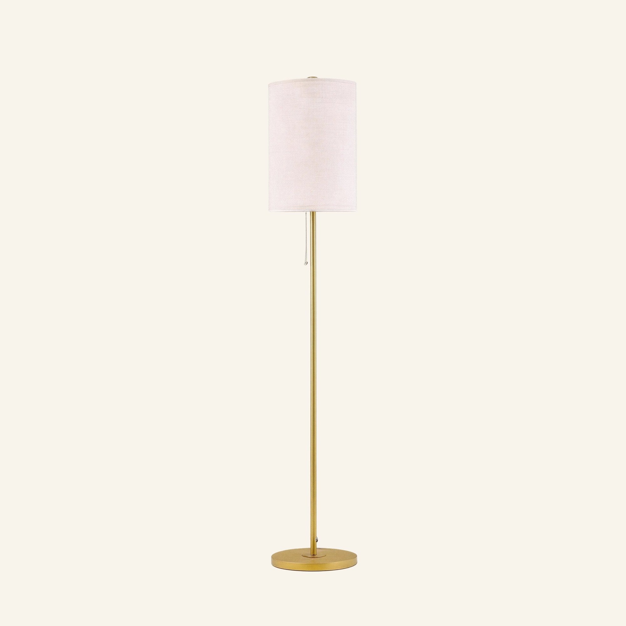 Slim floor lamp with cream linen drum shade on a slender brass-finished steel pole, round base, and pull-chain switch shown on cream backdrop.