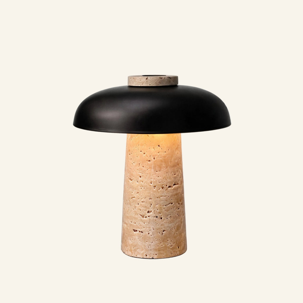 Cordless table lamp with tapered travertine column and matte black metal dome shade glowing softly from within on cream backdrop
