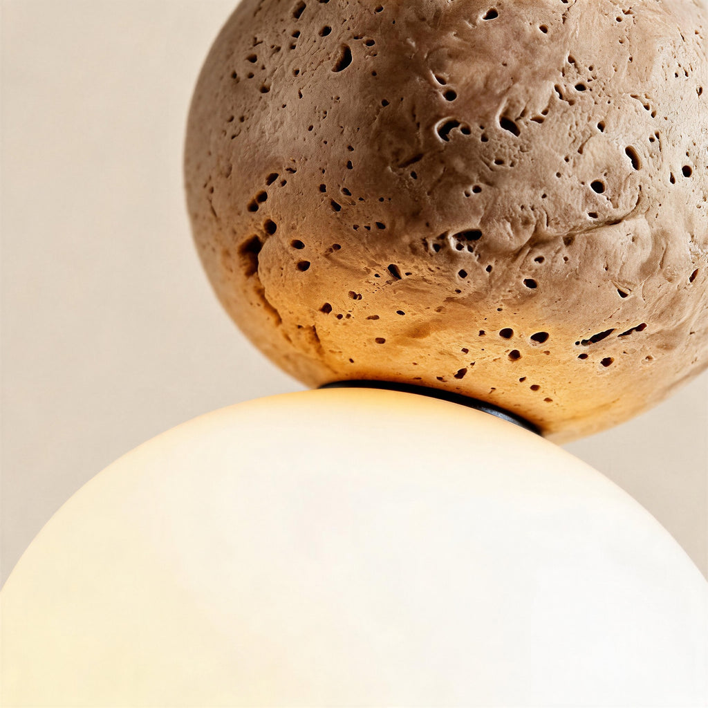 Close-up of porous travertine stone sphere joined to frosted white glass globe by a dark metal band, showing tactile pitted surface.