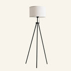 Tripod floor lamp with white linen drum shade on three slim black metal legs, with pull-chain cord, on cream background.