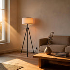 Tripod floor lamp with linen drum shade beside a wooden coffee table in a sunlit living room with beige sofa.