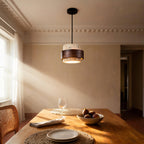 Stone and walnut pendant glowing above a wooden dining table set with figs, plates and a rattan chair in a sunlit room.