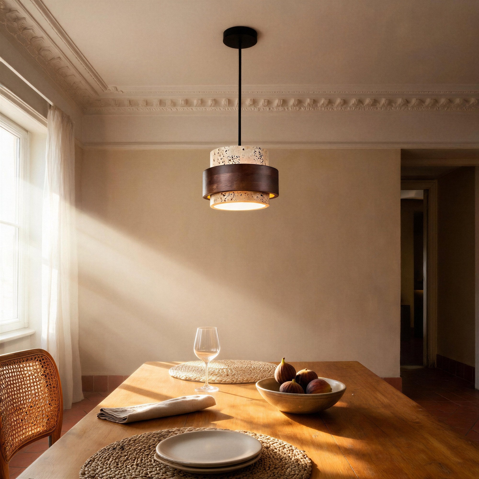 Stone and walnut pendant glowing above a wooden dining table set with figs, plates and a rattan chair in a sunlit room.